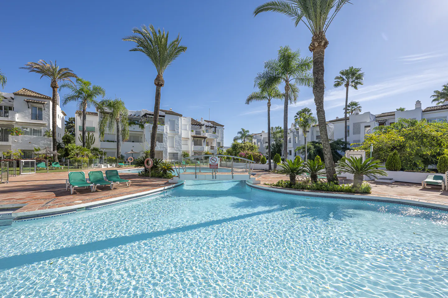 A bright, sunny view of a turquoise pool surrounded by palm trees and white buildings.
