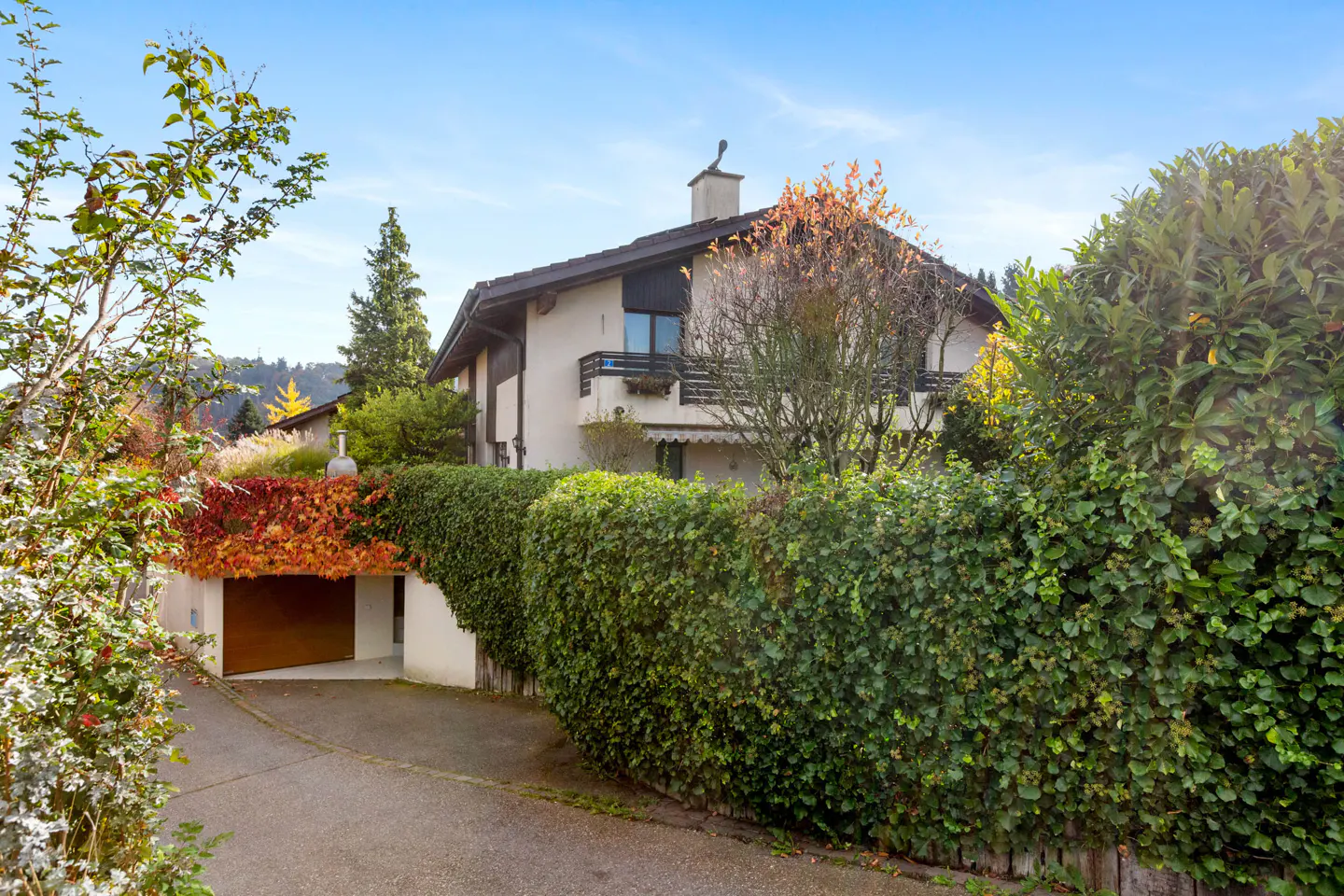 A two-story house with a brown roof, white walls, and a chimney, surrounded by green bushes and trees. A garage with a brown door is visible.