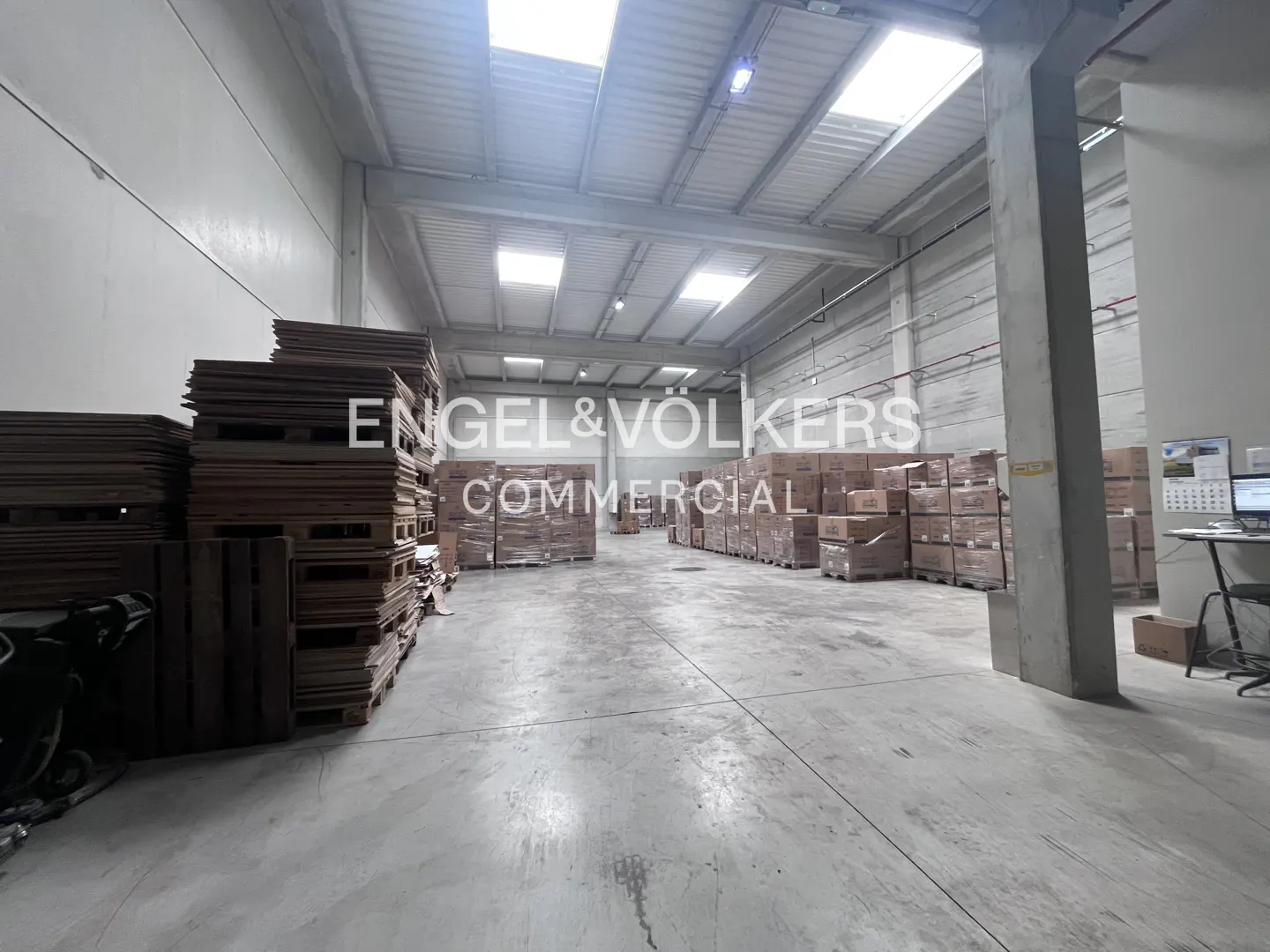 A large, bright warehouse with stacks of pallets and boxes. The Engel & Völkers logo is visible.