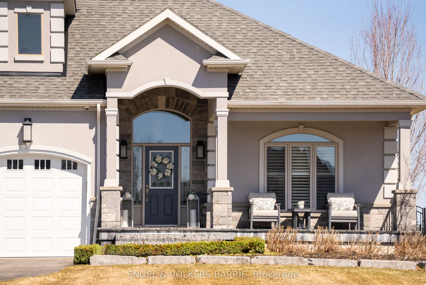 Beige house exterior with a gray roof, white garage door, and a blue front door with a wreath. Two chairs sit on the porch.