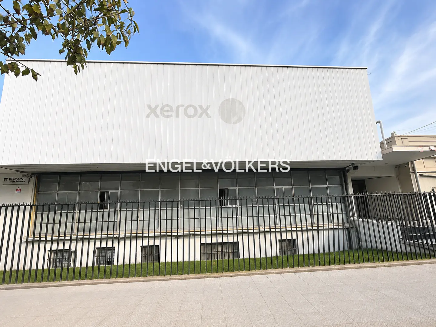 Exterior of a white commercial building with "Xerox" and "Engel & Volkers" logos, behind a black fence and green grass.