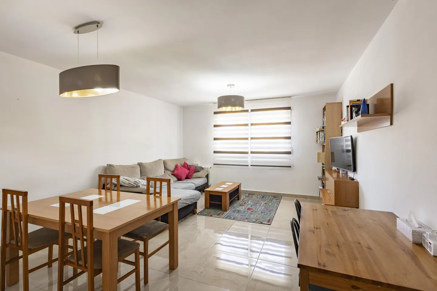 Bright living room with a wooden dining set, grey sectional sofa with pink pillows, coffee table, and wall-mounted TV.