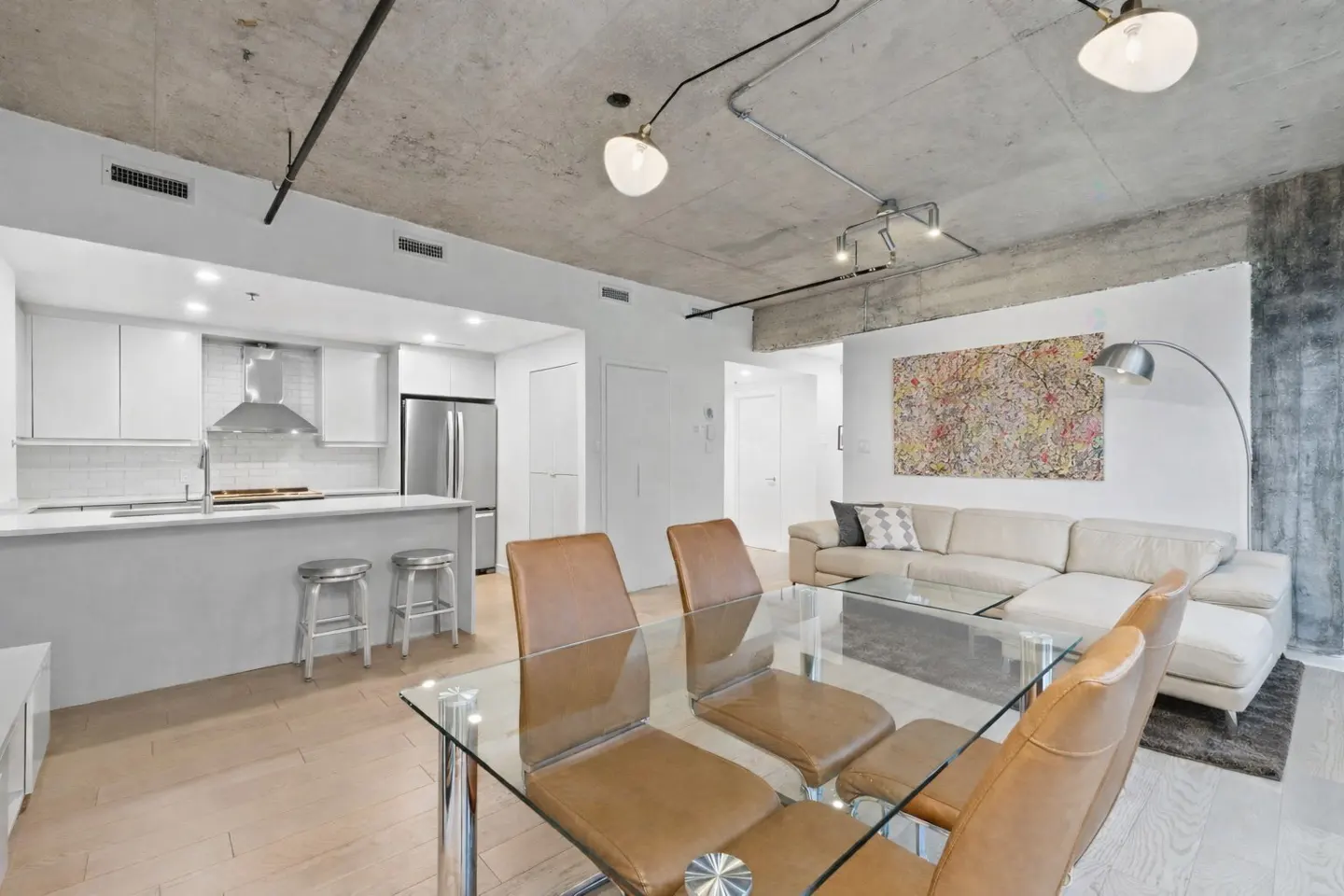 Open-concept living space with a modern kitchen, glass dining table with brown chairs, and a cream-colored sofa. Concrete ceiling with exposed pipes.