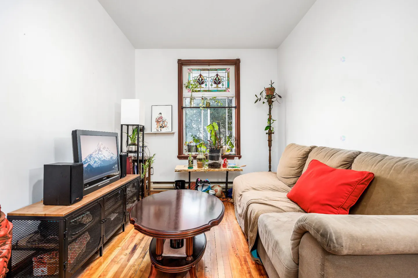 Living room with white walls, wood floors, and a brown sofa with a red pillow. A TV sits on a metal and wood stand. A stained glass window is above a table with plants.
