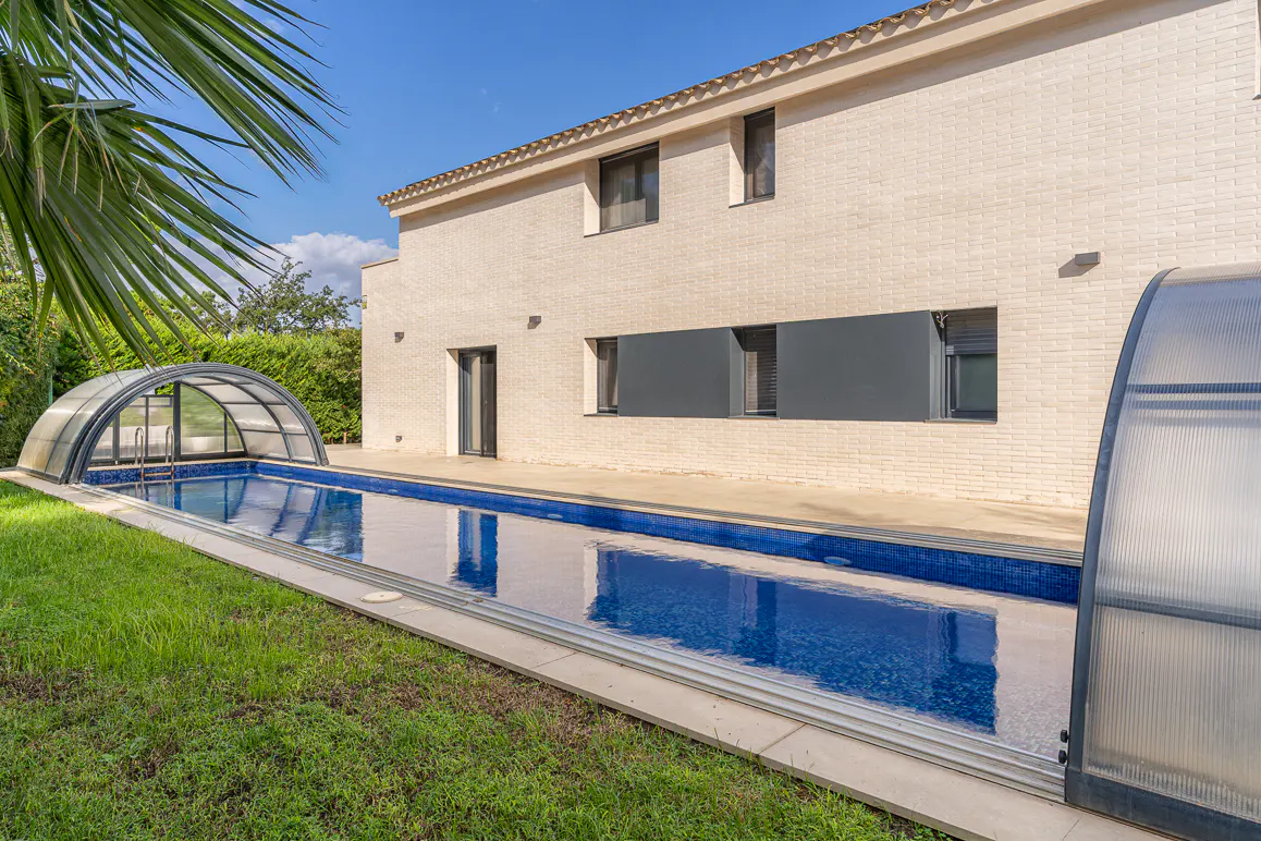 A long, blue tiled pool with a clear cover sits beside a two-story, beige brick house with dark windows and a green lawn.