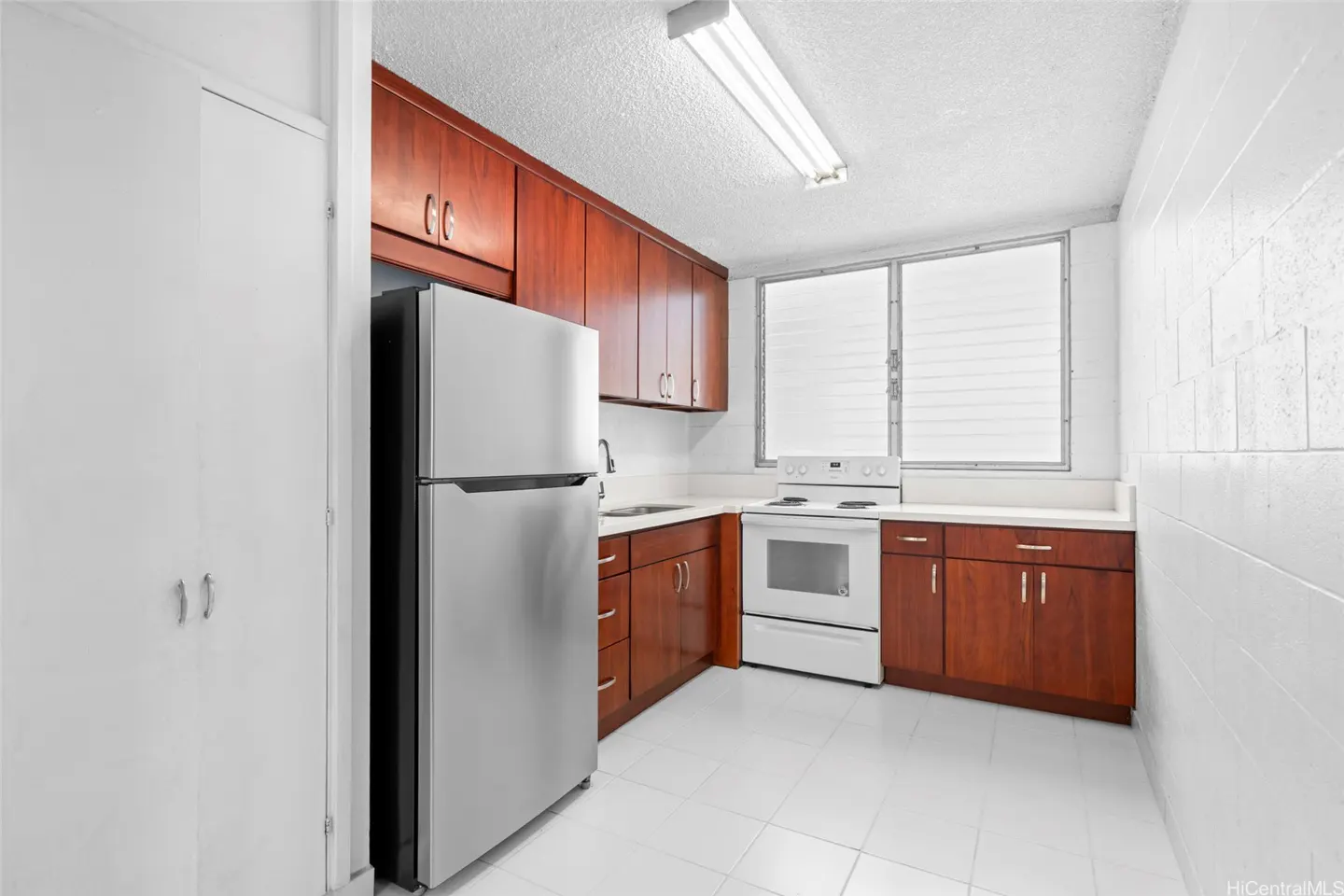 A kitchen with white tile floors, white walls, and dark wood cabinets. A stainless steel refrigerator is on the left. A white stove is near the window.