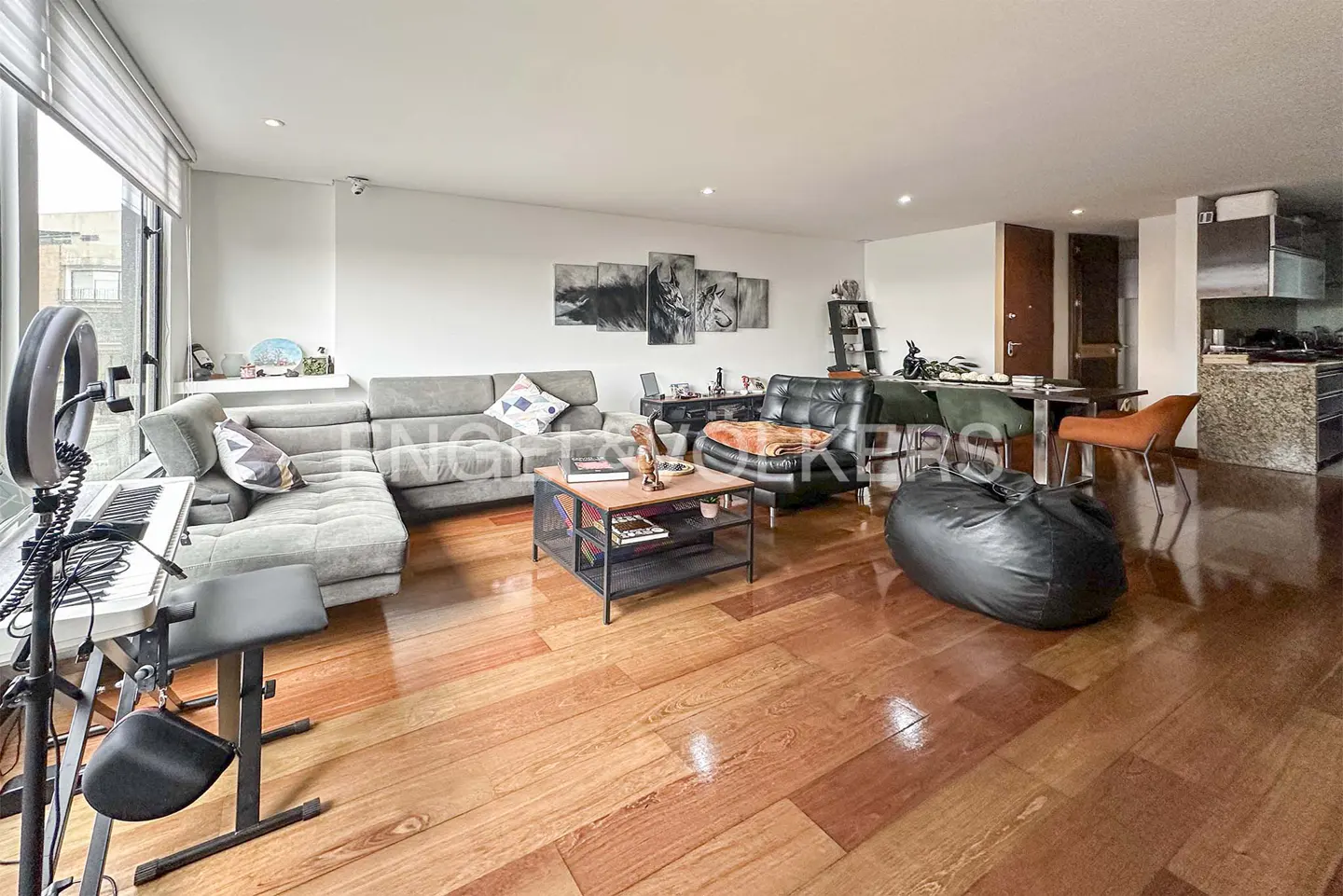 Bright living room with hardwood floors, grey sectional sofa, black leather chair, and a keyboard with a ring light.