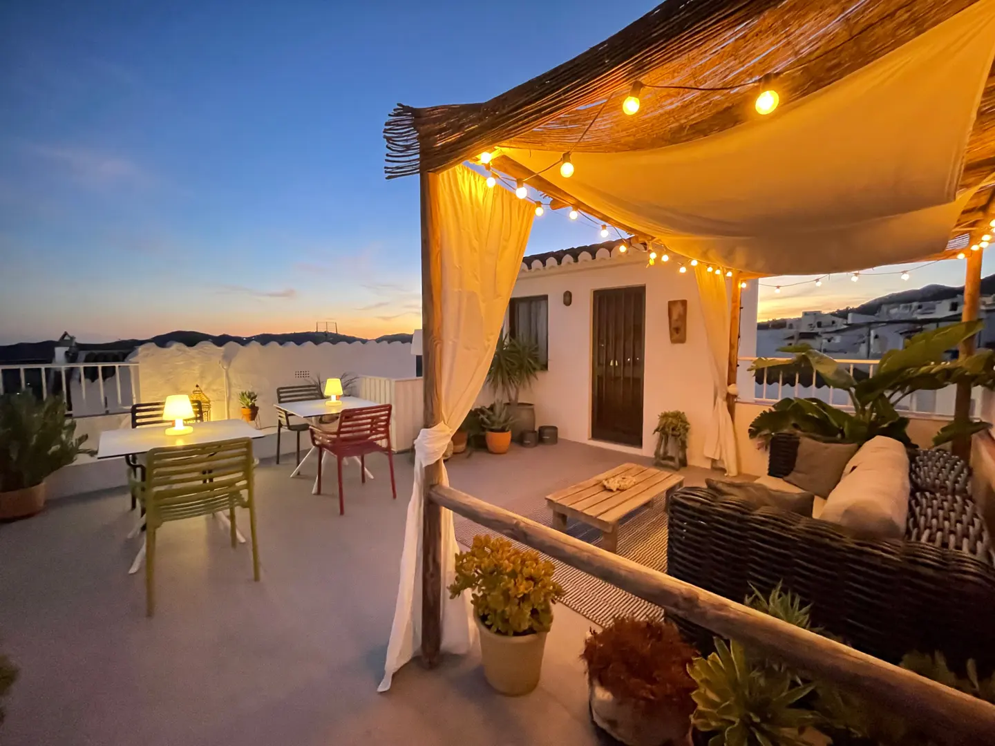 Rooftop patio at dusk with string lights, tables, chairs, and a wicker sofa. Plants add greenery to the outdoor space.