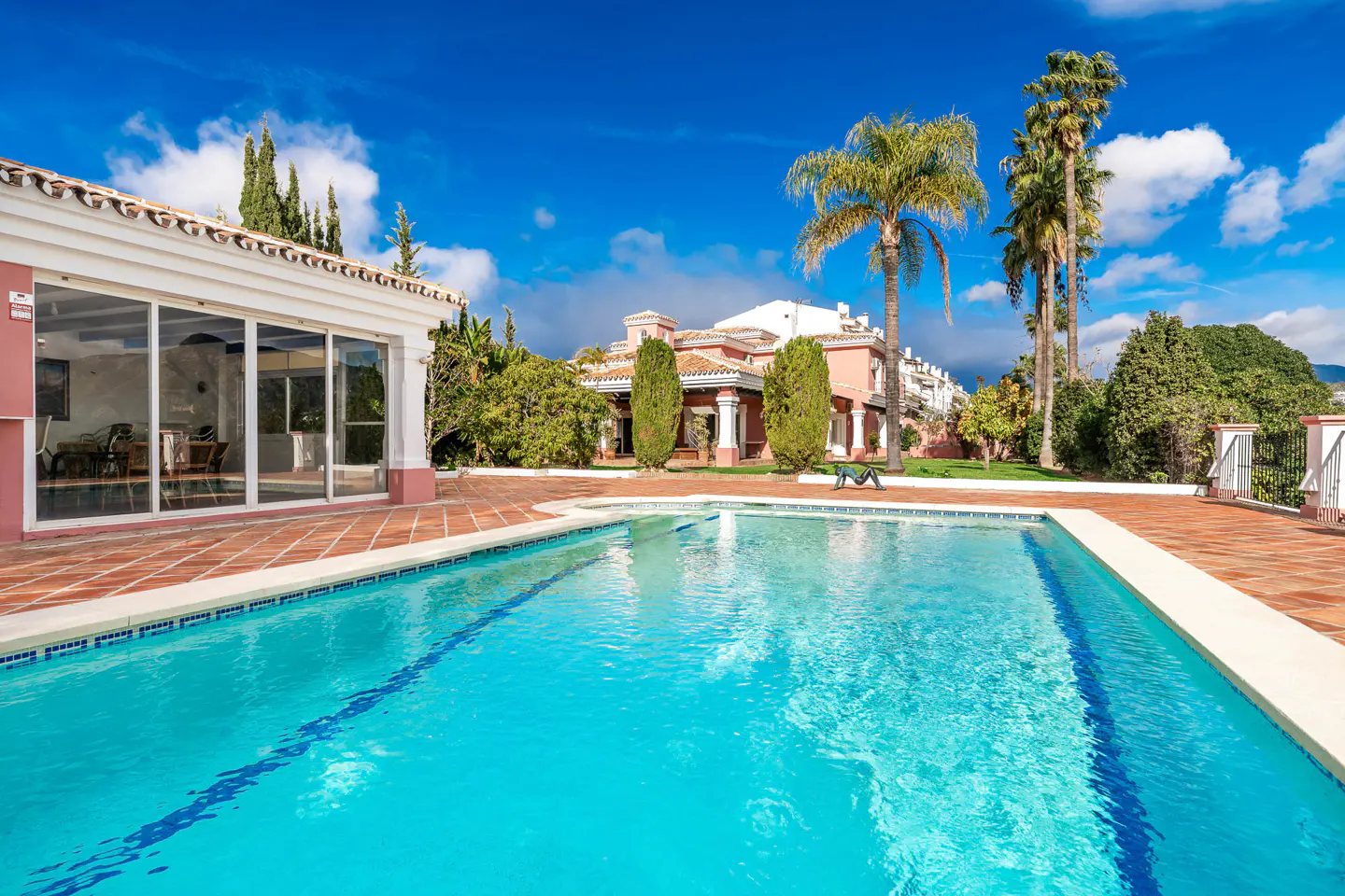 A turquoise swimming pool in front of a pink house with palm trees under a blue sky.