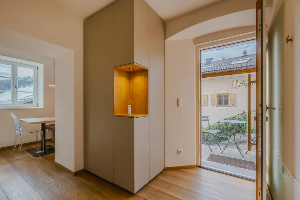 Interior view of a room with wood floors, a built-in cabinet with a lit niche, and an open door to a patio with outdoor furniture.