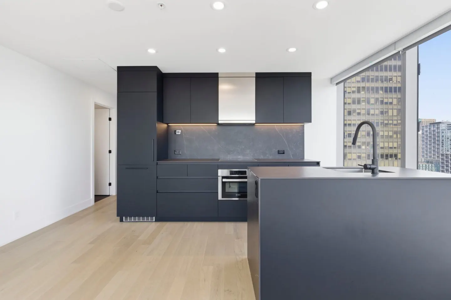 Modern kitchen with dark gray cabinets, stainless steel appliances, and a large island with a black faucet, city view through the window.