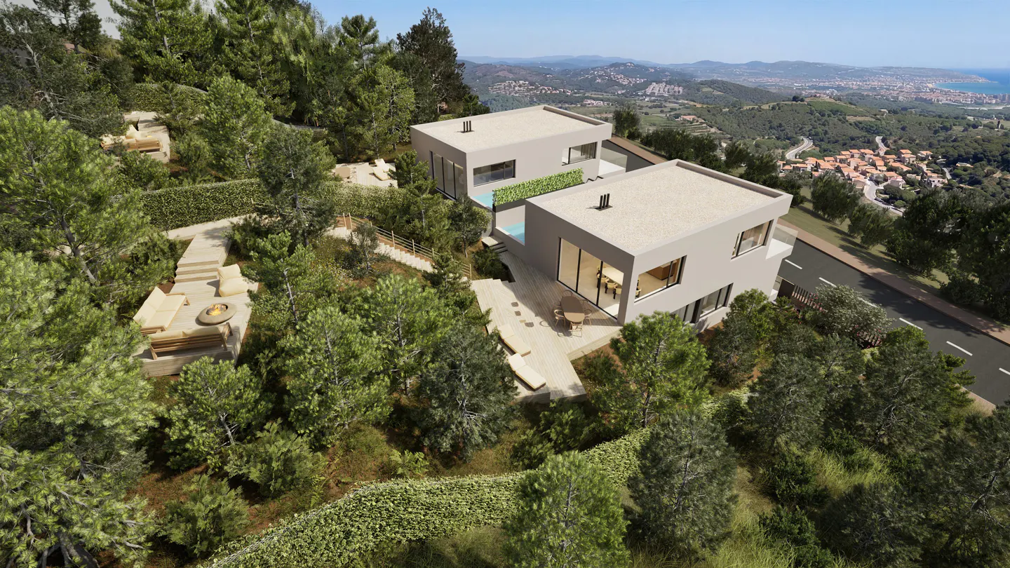 Aerial view of two modern, gray, flat-roofed houses nestled on a wooded hillside with a distant coastal town in the background.