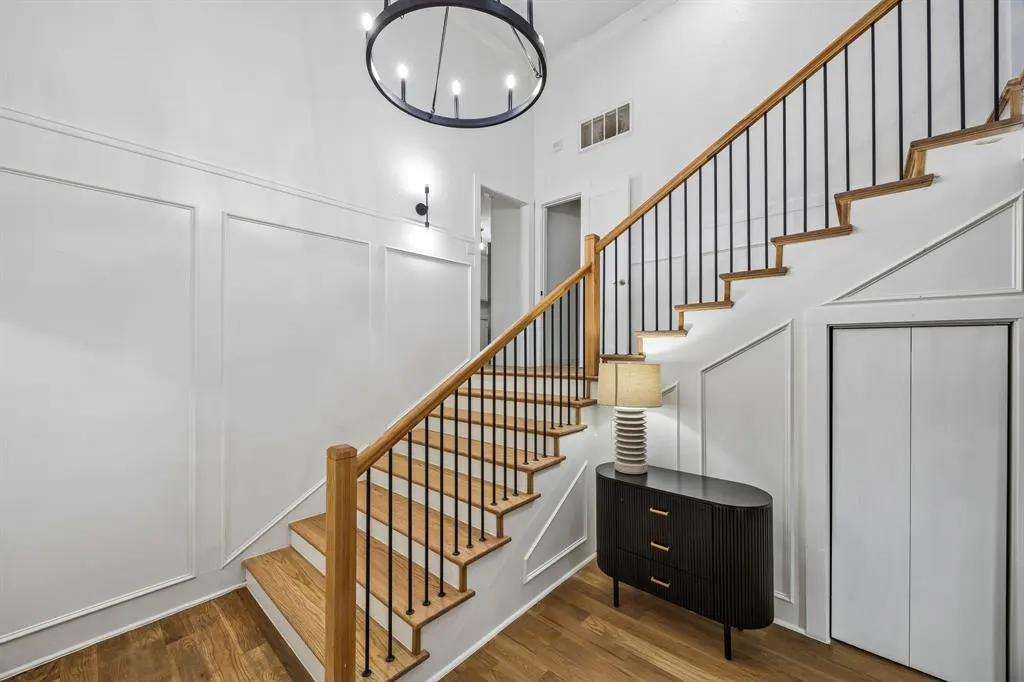 Bright foyer with wood floors, white walls, and a staircase with wood steps and black railings. A black cabinet and lamp sit near a closet.