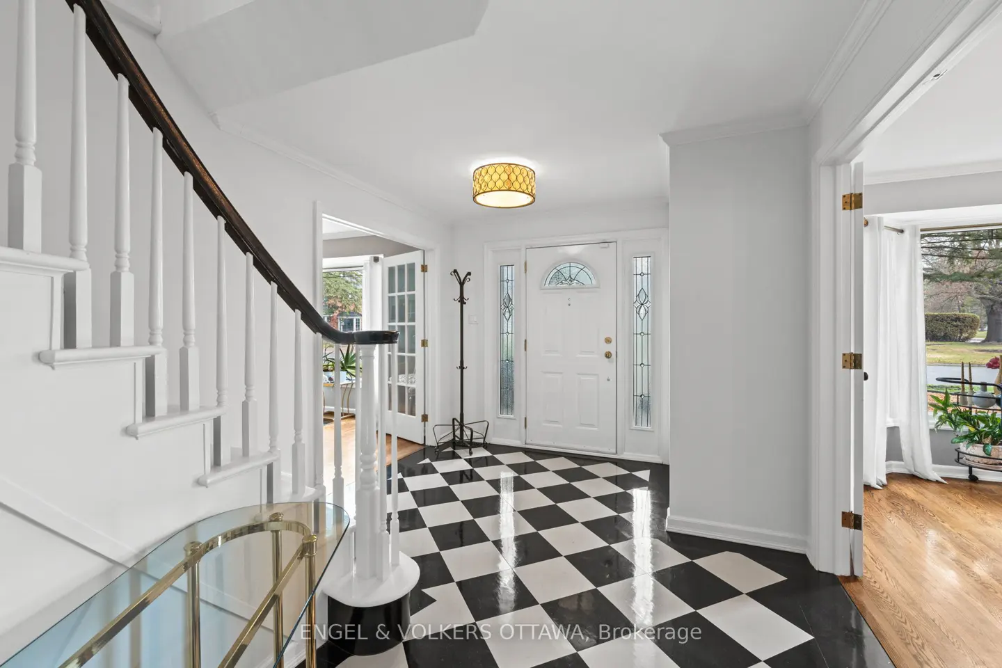 Bright foyer with black and white checkered floor, white walls, and staircase. A white front door with glass panels is centered.