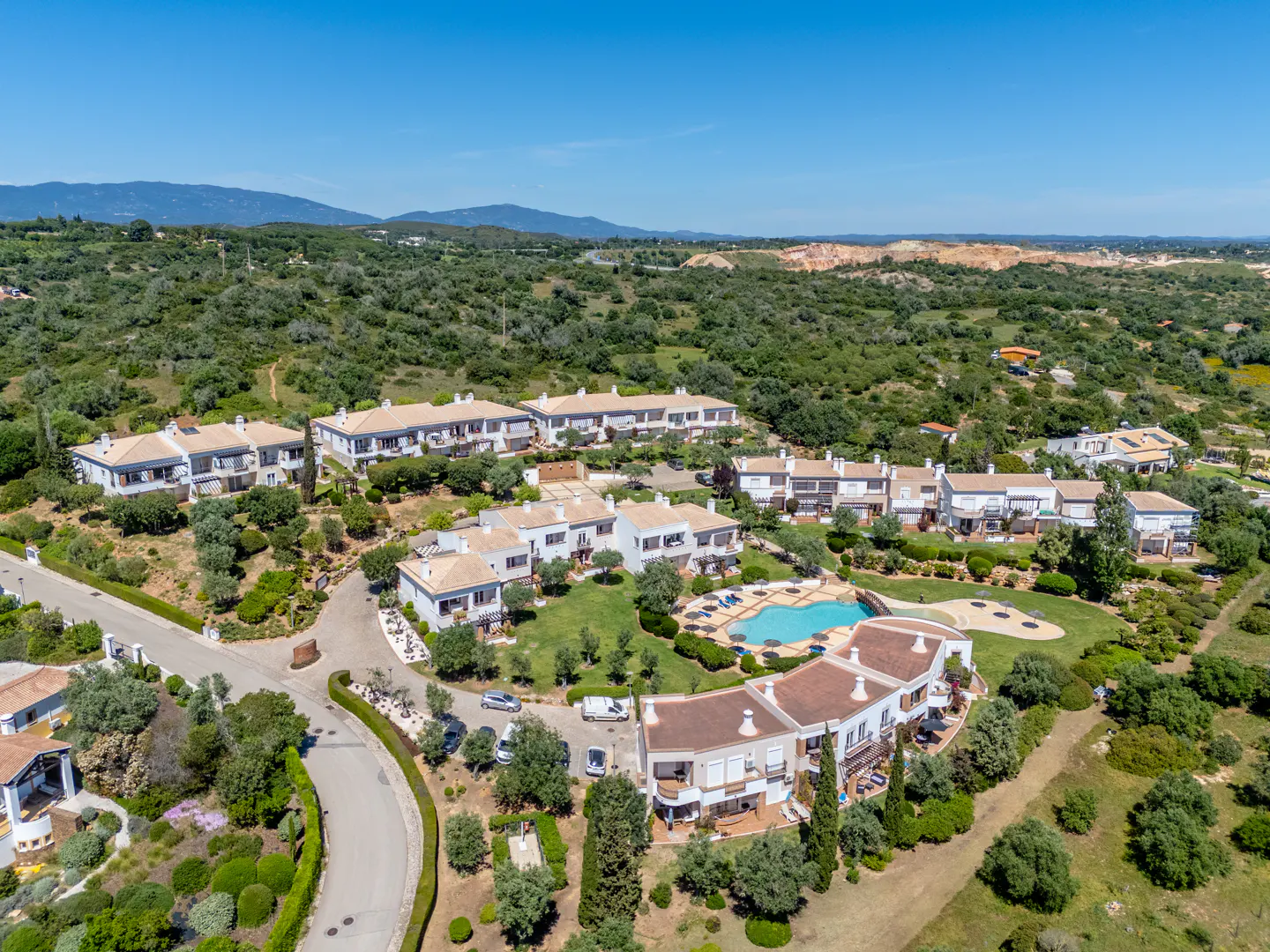 Aerial view of a residential complex with white buildings, brown roofs, a pool, and green landscaping under a blue sky.