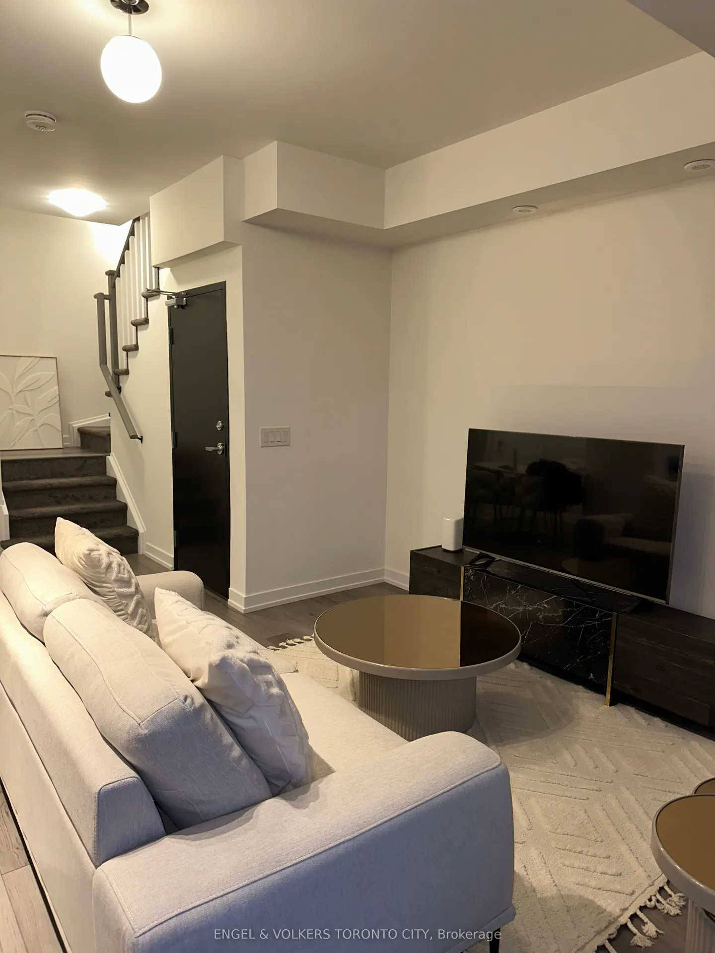 Living room with a white sofa, round coffee table, TV, and stairs leading up. The walls are white, and there is a black door.