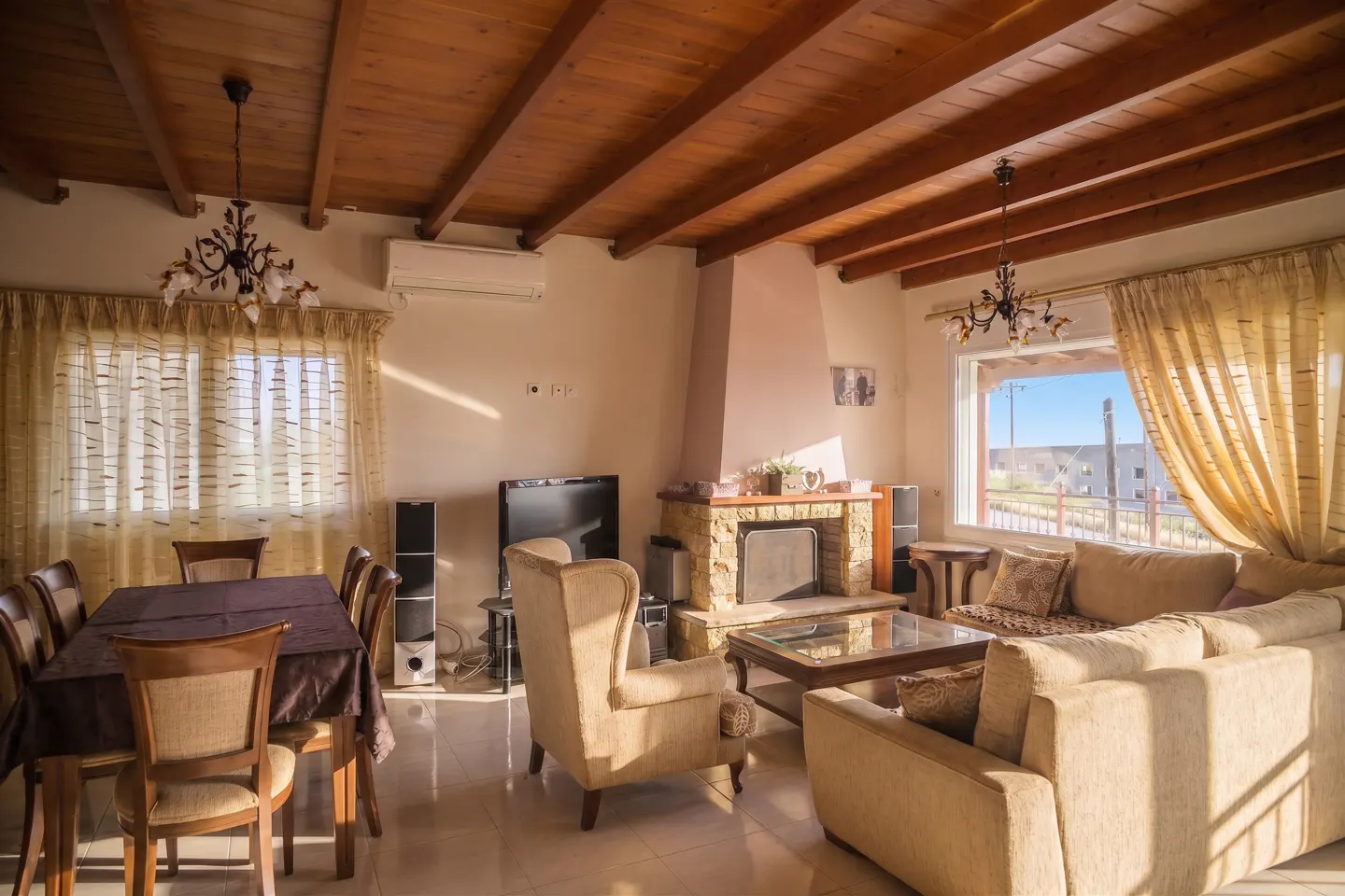 Living room with wood-beamed ceiling, stone fireplace, beige sofa, dining table, and large window.