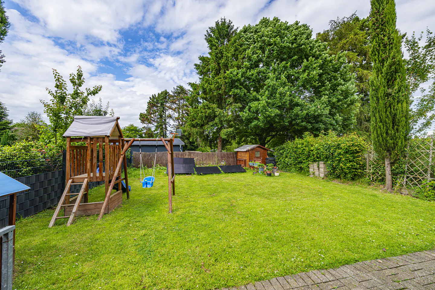 Spacious green backyard with a wooden playset, blue swing, small shed, and mature trees under a cloudy blue sky.