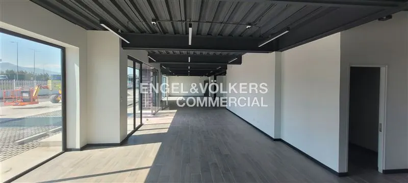 Empty commercial space with gray wood floors, white walls, and a black ceiling with fluorescent lights. Large windows offer an outside view.