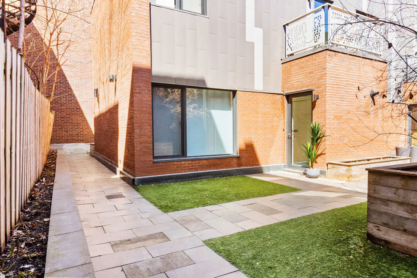 Brick building exterior with a small yard. A stone path leads to a door with a potted plant. There is a small patch of green grass.