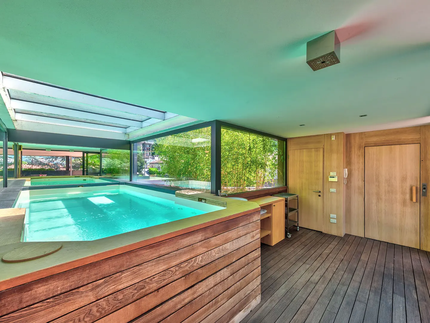 Indoor pool area with wood floors, a glass ceiling, and a large window with bamboo outside. Two wooden doors are on the right.