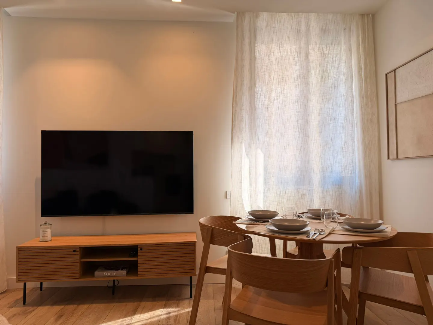 Living room with a TV above a wooden cabinet, a round dining table set for four, and light-colored curtains.