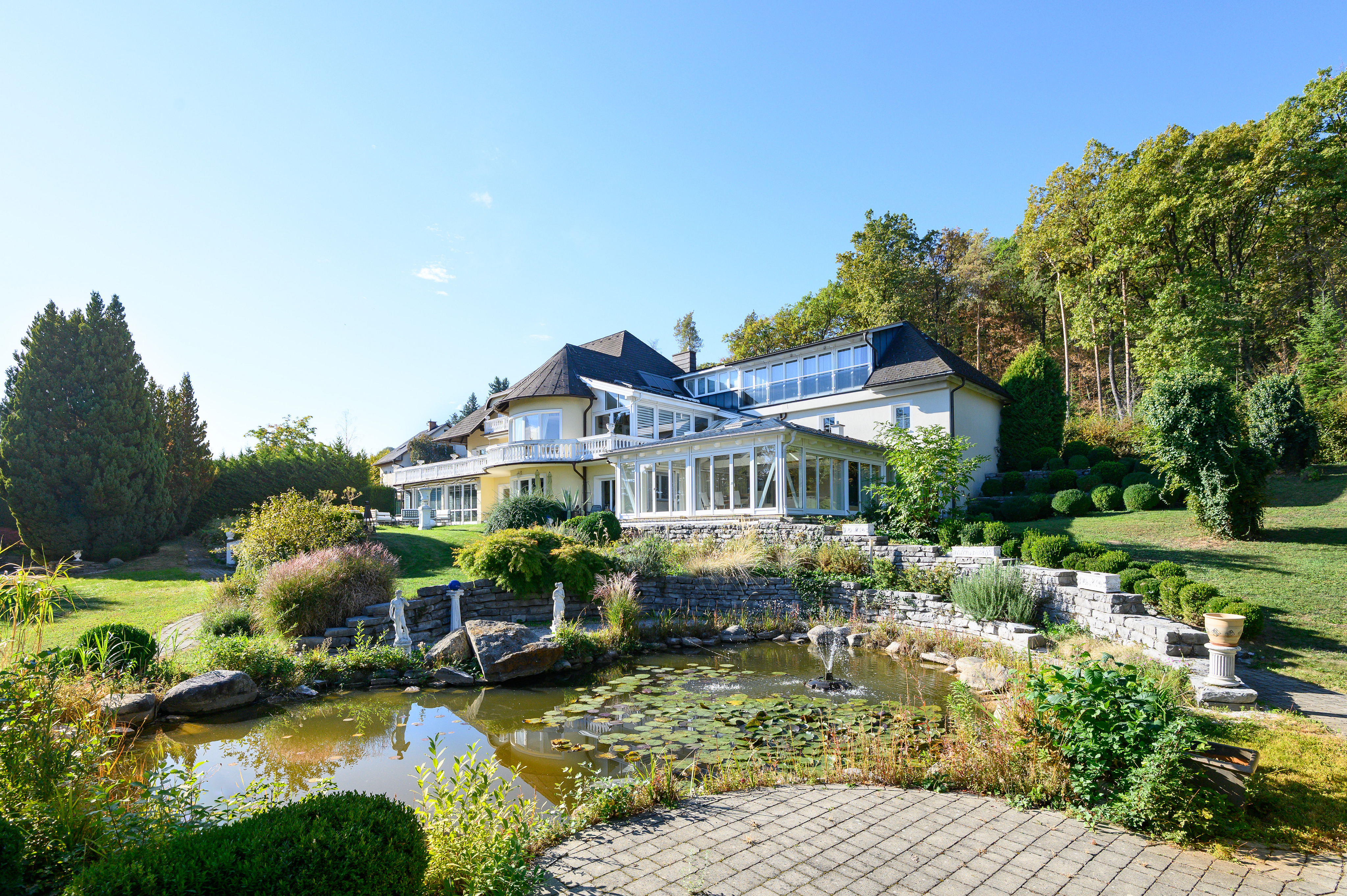 Large cream-colored mansion with a sunroom, overlooking a garden pond with a fountain and stone retaining walls. [AI-generated text]