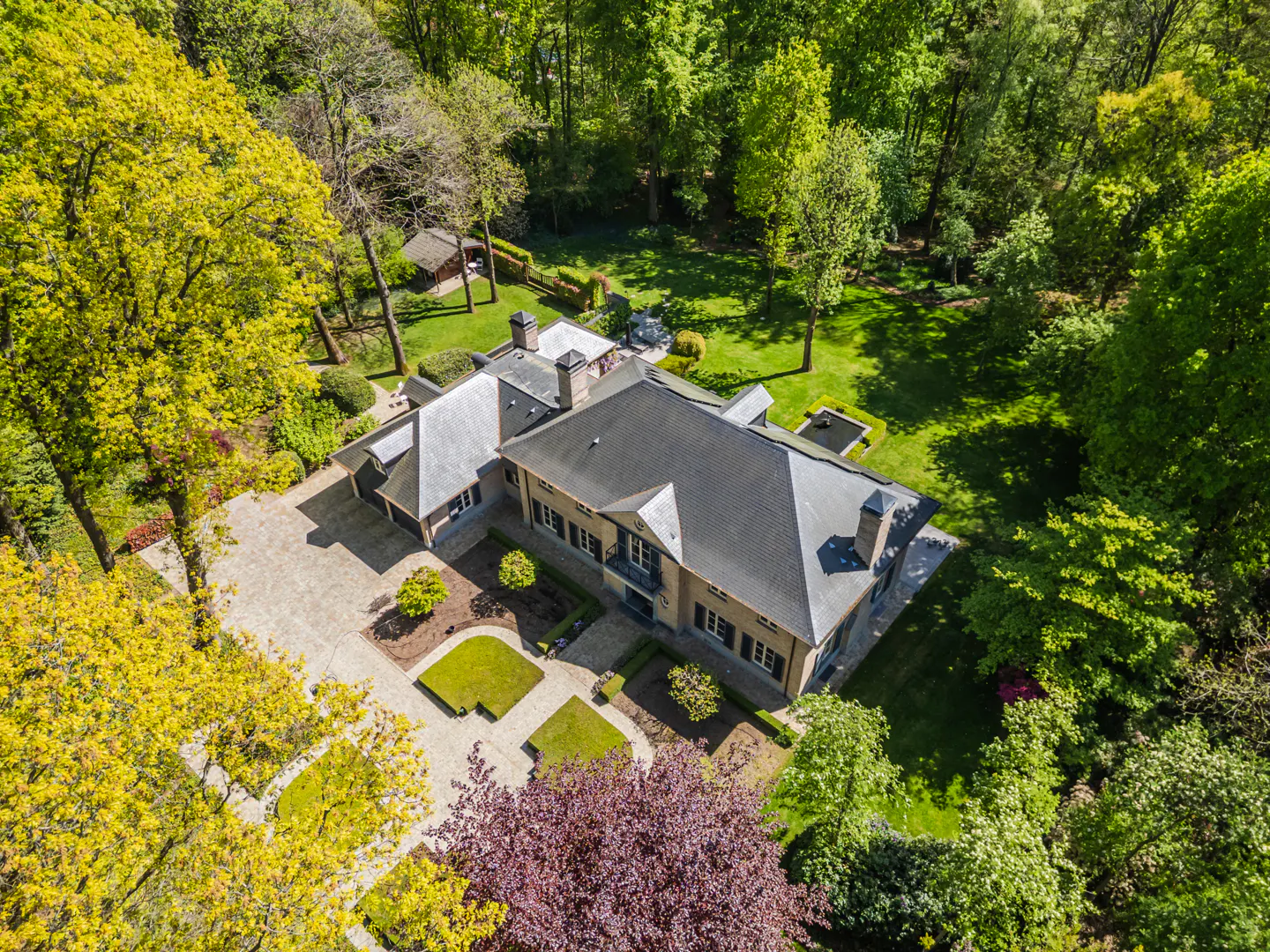 Aerial view of a large, grey-roofed house surrounded by green trees and manicured gardens on a sunny day.