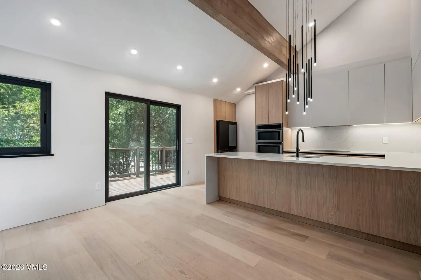 A modern kitchen with light wood floors, white walls, and a wood island with a white countertop. Black framed windows and sliding doors look out to trees.