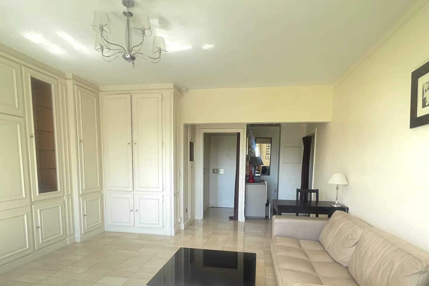 Living room with beige walls, tile floors, and built-in cabinets. A beige sofa faces a black coffee table. A chandelier hangs from the ceiling.
