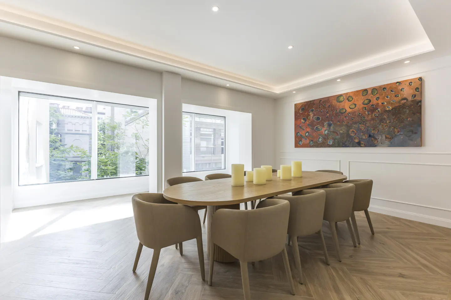 Bright dining room with a long wooden table, beige chairs, and white candles. Large windows overlook greenery, and an abstract painting hangs on the wall.