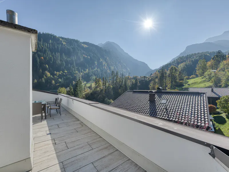 Balcony view of mountains. Outdoor table and chairs on a stone patio with a mountain range and blue sky in the background.