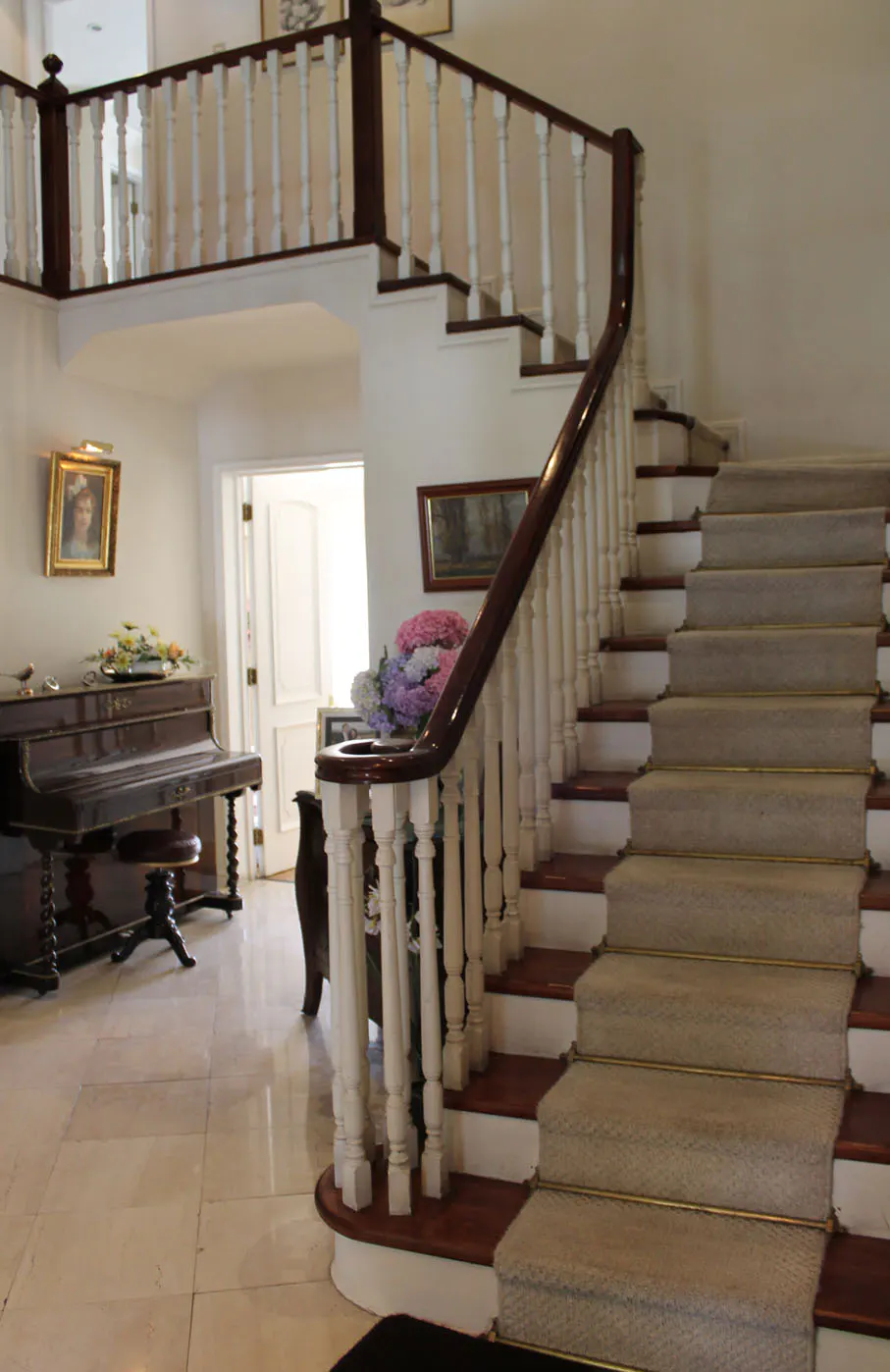 Grand foyer with a curved staircase, white balusters, and a dark wood railing. A piano sits to the left, with artwork and a doorway in the background.
