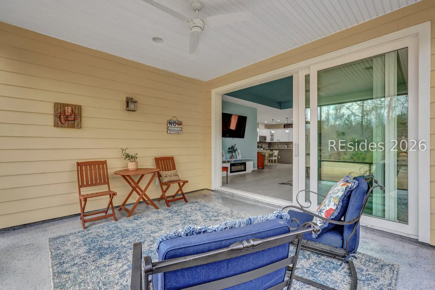 Covered patio with blue rug, rocking chair, and table with chairs. Sliding glass doors open to a modern kitchen. Yellow siding and white ceiling.