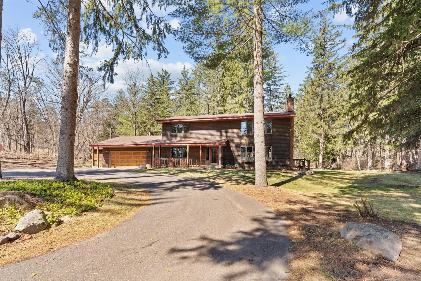 Two-story brown house with a front porch and attached garage, surrounded by trees and a driveway.