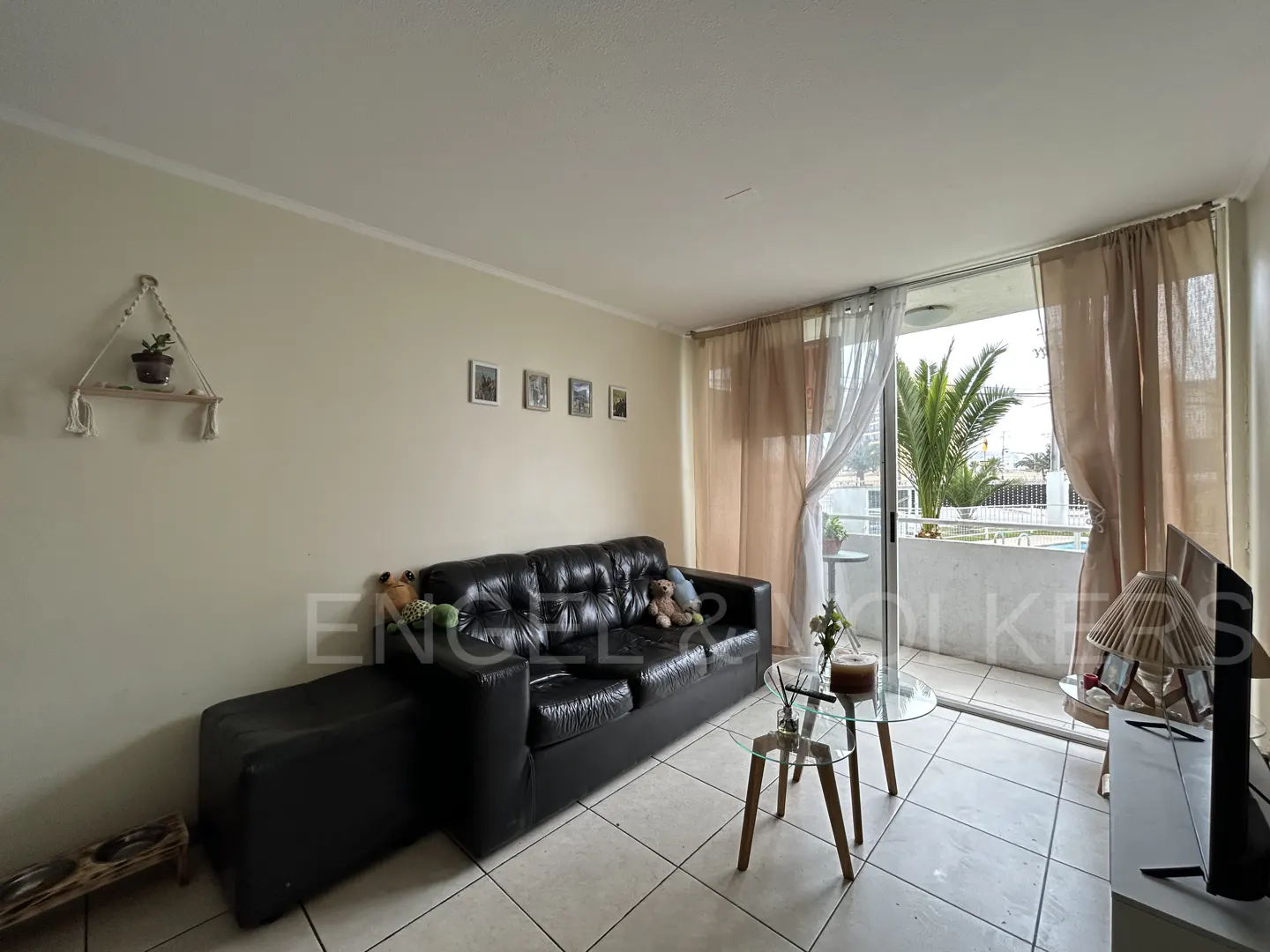 Living room with a black leather sofa, glass coffee tables, and a sliding door leading to a balcony with palm trees.