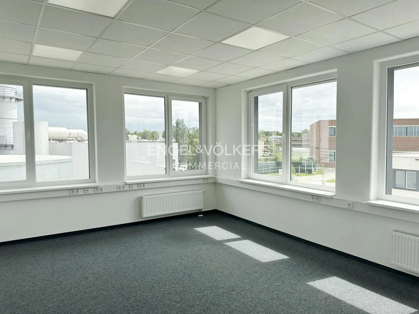 Bright, empty office space with gray carpet, white walls, and large windows showing an exterior view.