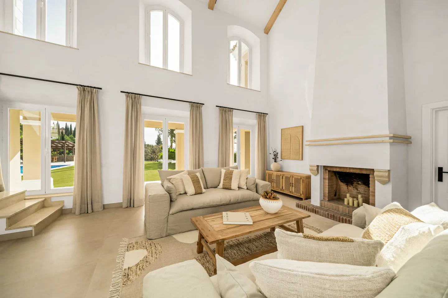 Bright living room with high ceilings, white walls, and large windows. A beige sofa and wooden coffee table sit on a patterned rug. A fireplace and outdoor view are visible.