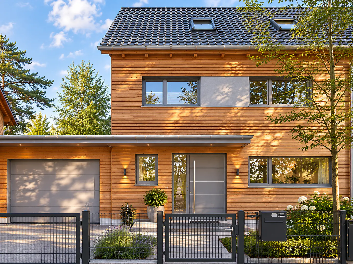 Two-story house with wood siding, gray trim, and a gray garage door. A black metal fence surrounds the front yard.
