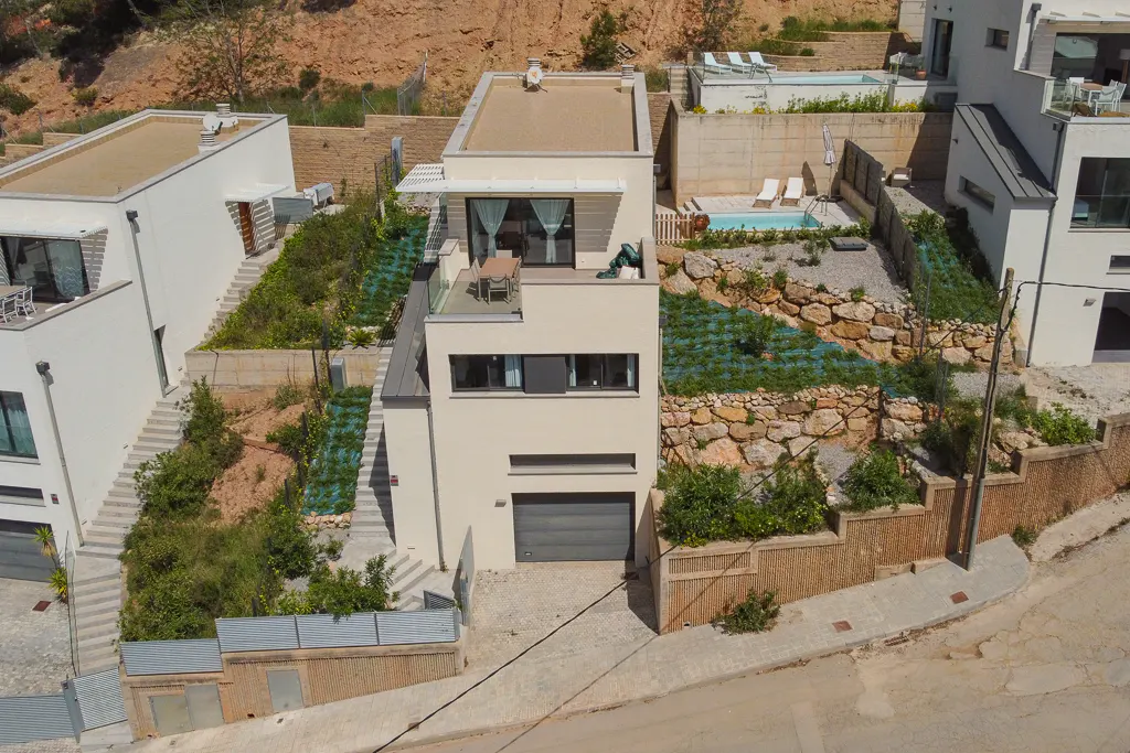 Aerial view of a modern, three-story white house with a gray garage door, surrounded by tiered gardens and neighboring homes on a sunny day.