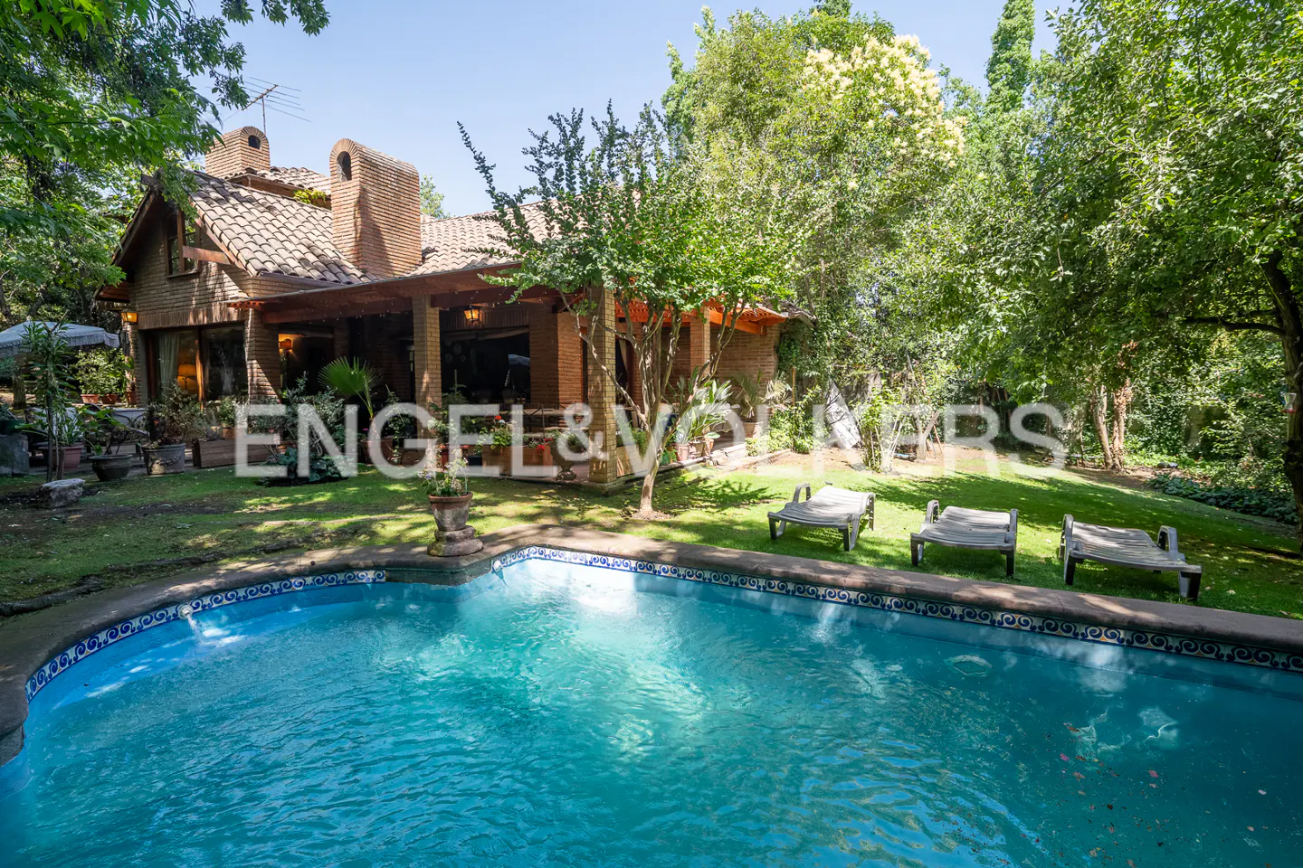 A brick house with a red tile roof overlooks a blue swimming pool and green lawn with lounge chairs.