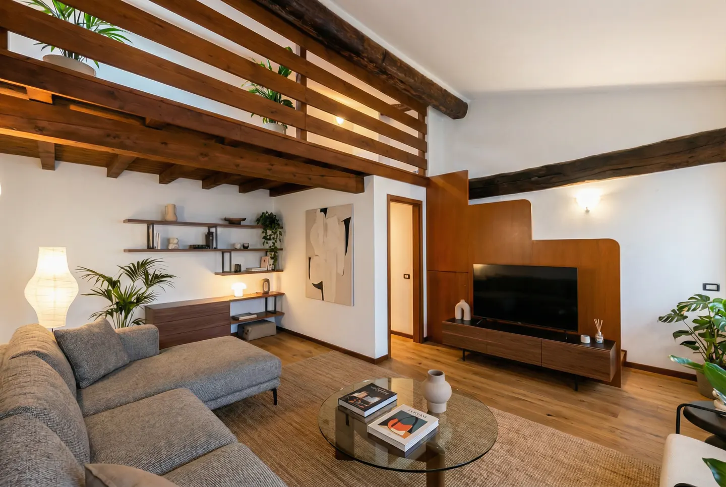 A modern living room with a gray sectional sofa, glass coffee table, and wooden beams on the ceiling. Shelves and a TV stand are also visible.