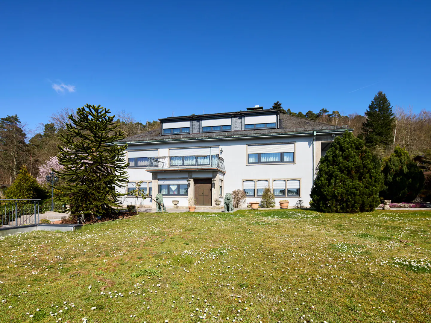 A two-story white house with a gray roof sits on a green lawn with white flowers under a blue sky.