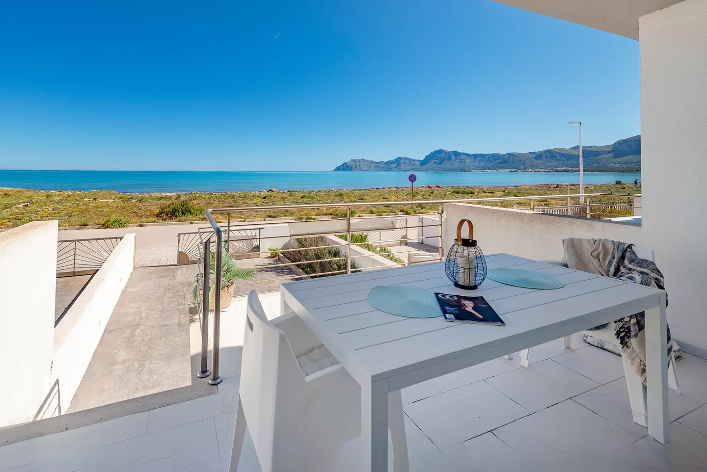 Balcony view of the ocean with a white table, chairs, lantern, and magazine. Mountains are visible in the distance.