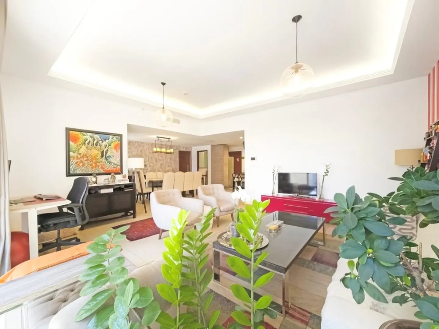 Bright, open-plan living room with white walls, beige sofas, and a black coffee table. A red cabinet sits under a TV. Dining area visible in the background.