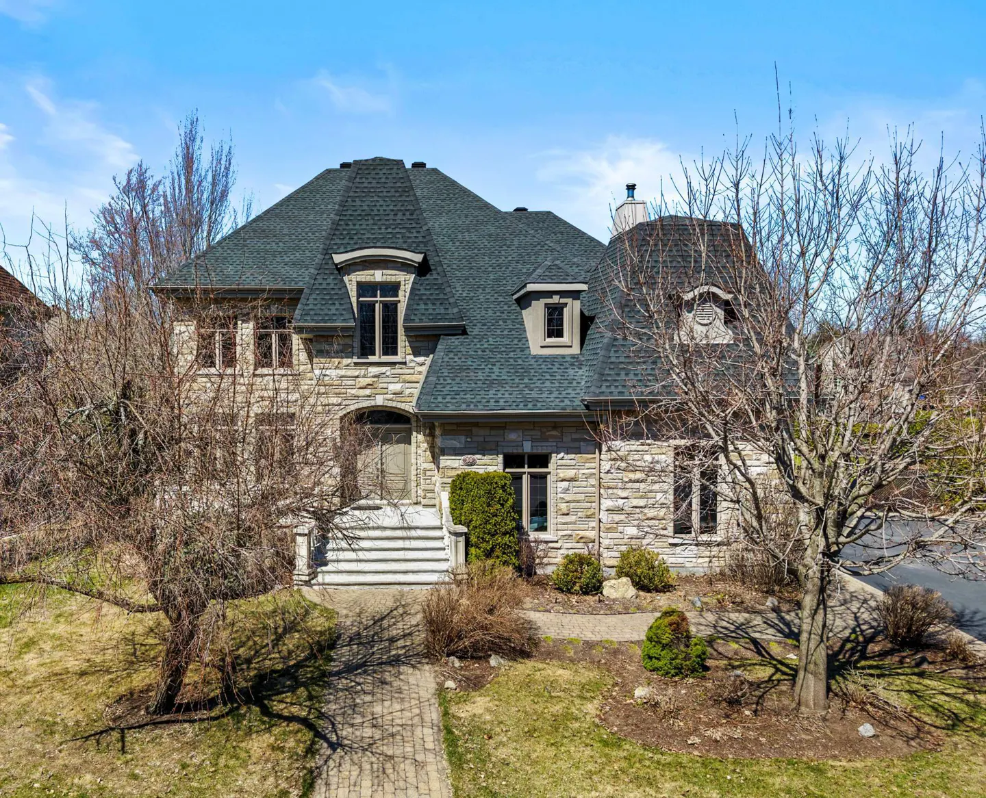Two-story stone house with a gray roof, dormer windows, and bare trees in the front yard.