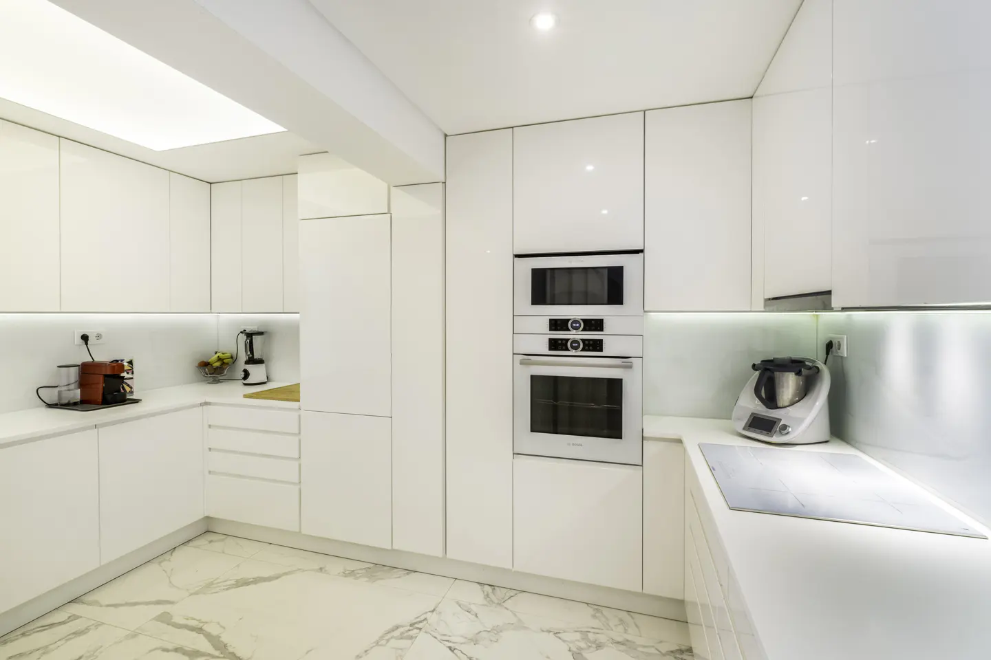 A modern, all-white kitchen with marble floors, built-in appliances, and under-cabinet lighting.
