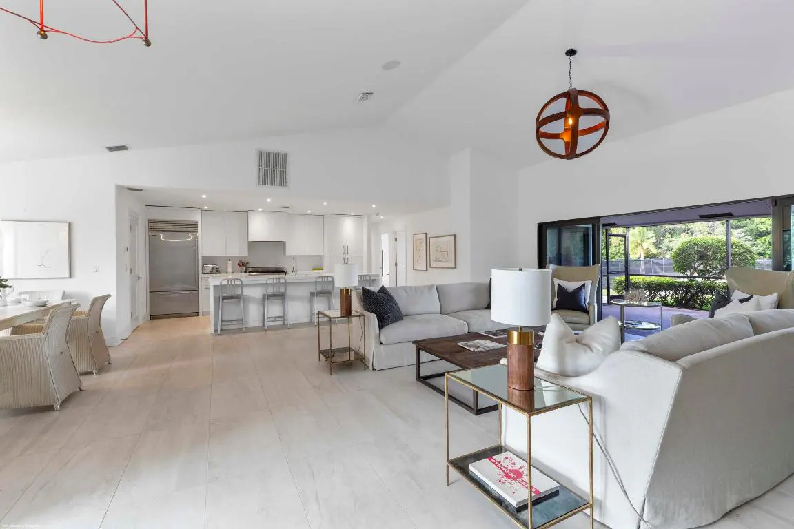A bright, open-concept living space with white walls, light wood floors, and modern furniture. A kitchen is visible in the background.