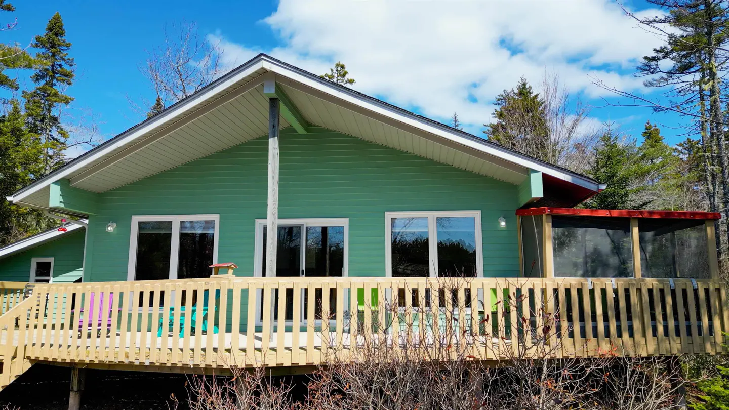 Exterior of a green house with a wooden deck and colorful chairs, surrounded by trees under a blue sky.