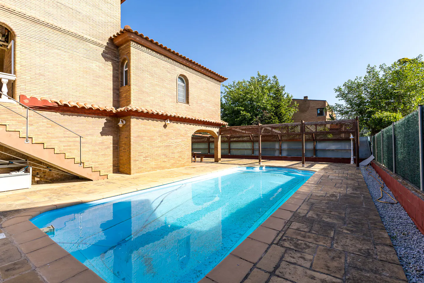 Backyard view of a brick house with a blue swimming pool, a wooden pergola, and a stone patio on a sunny day.